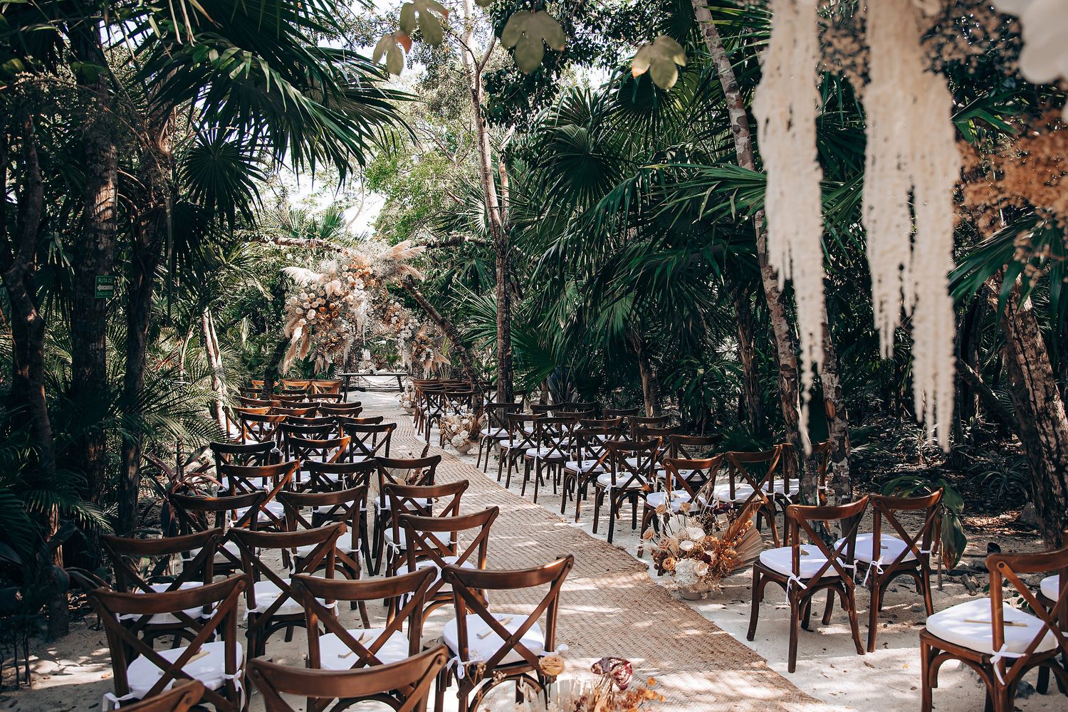 A rustic outdoor wedding venue with wooden cross-back chairs arranged in rows beneath towering palm trees and Spanish moss.