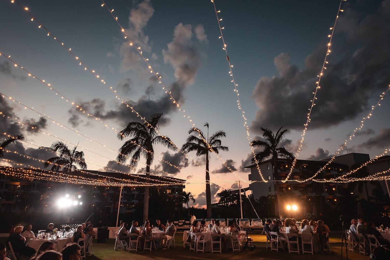 Outdoor wedding reception under string lights and palm trees at sunset with dramatic clouds.