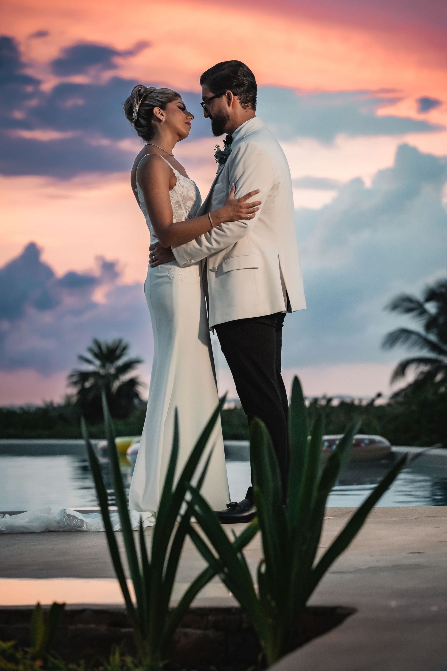 Romantic wedding portrait with tropical plants and pink sunset sky over water.