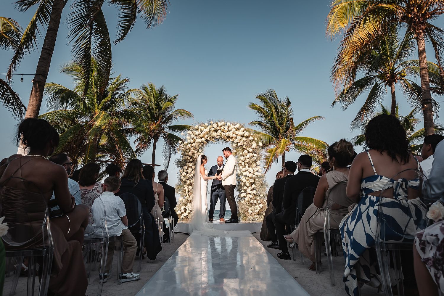 Wedding ceremony on white aisle runner with floral arch and palm trees during golden hour.