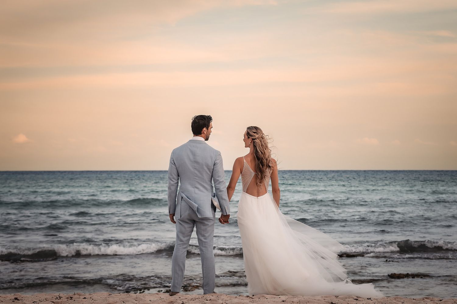 Newlyweds walk along a sandy beach at sunset with ocean waves in the background.