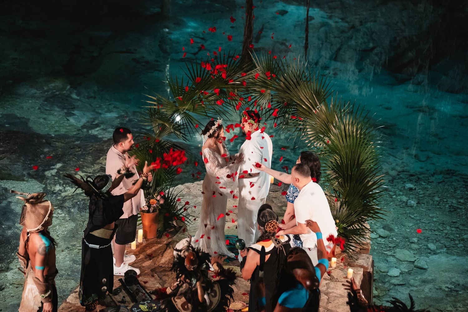 A group of people celebrate at a romantic nighttime beach wedding ceremony under palm trees and red lighting.