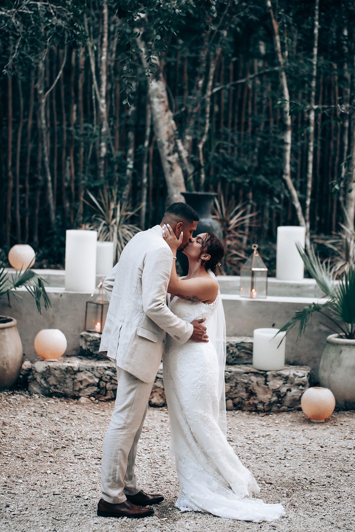 A romantic newlywed couple shares their first kiss at a forest wedding decorated with white pillars and peach balloons.