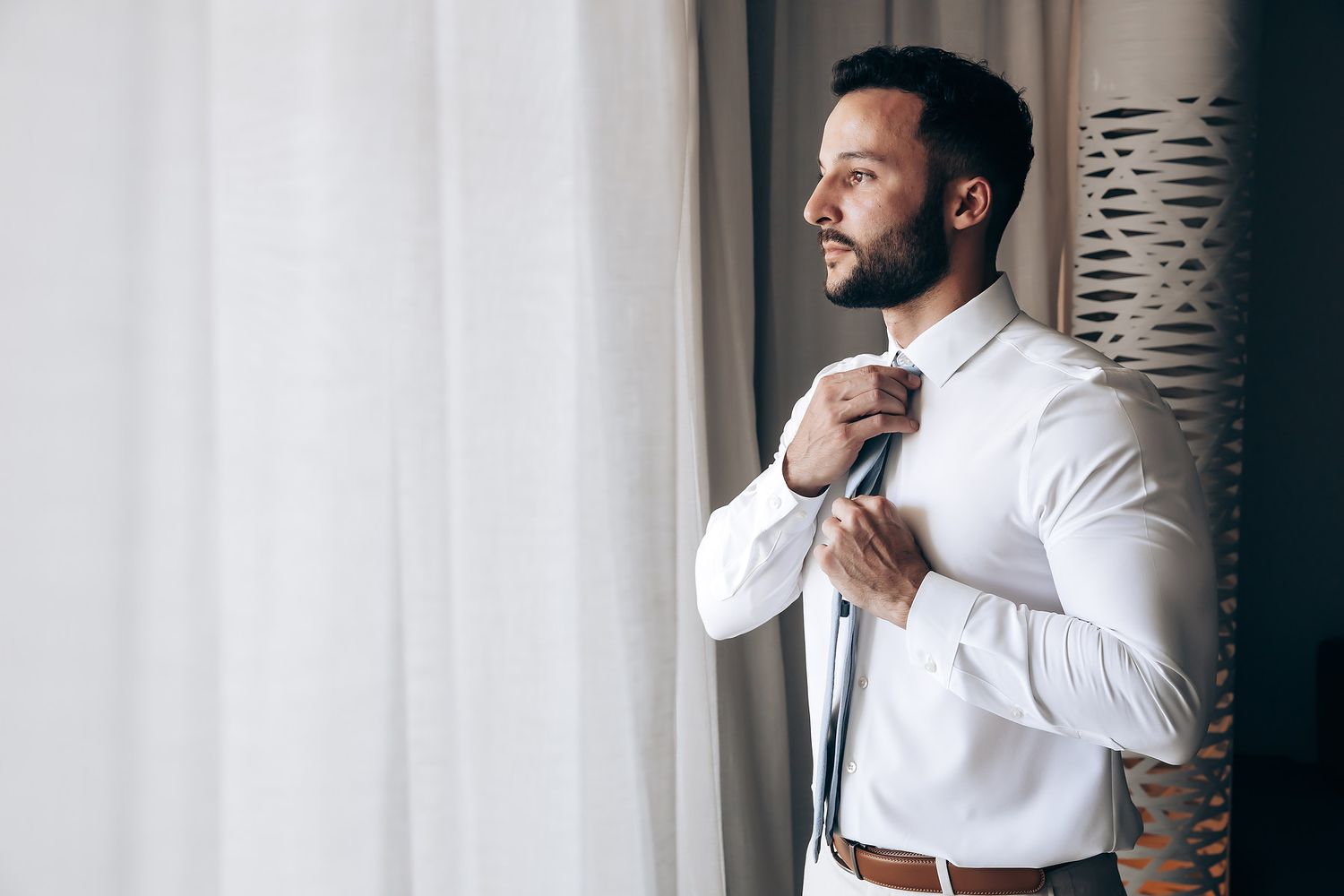 Groom getting ready in a crisp white shirt while adjusting cuffs near a window.