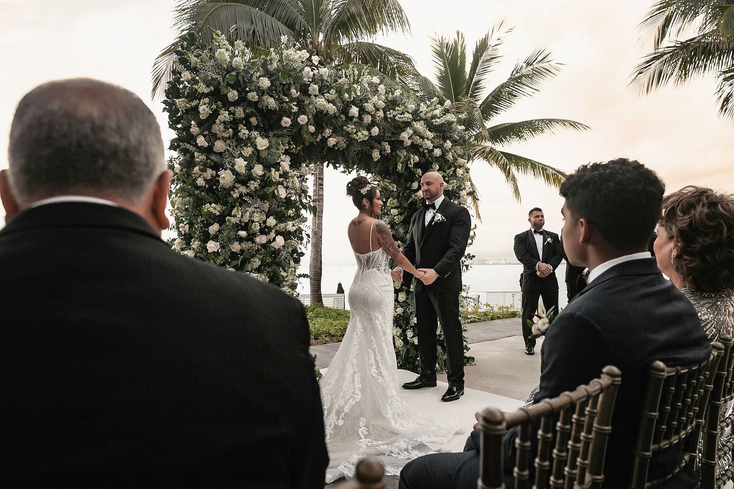 A wedding ceremony takes place outdoors at sunset with palm trees and ocean views as guests watch the couple exchange vows.