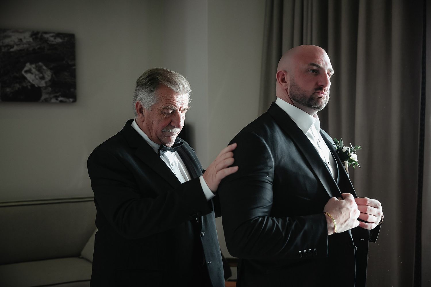 Two people in formal attire stand near a window in a dimly lit room.