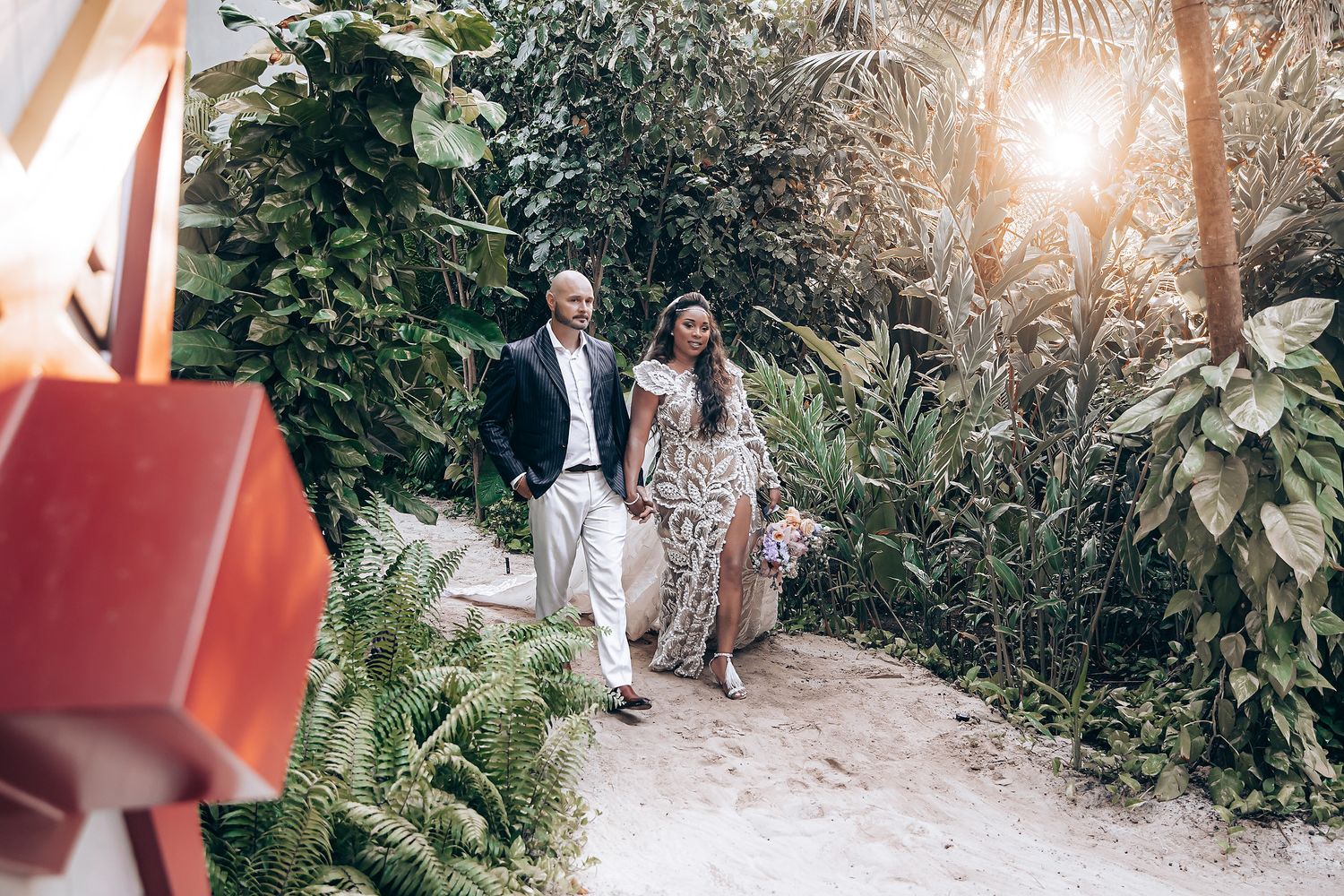 A couple walks along a garden path surrounded by green foliage as sunlight filters through the trees.