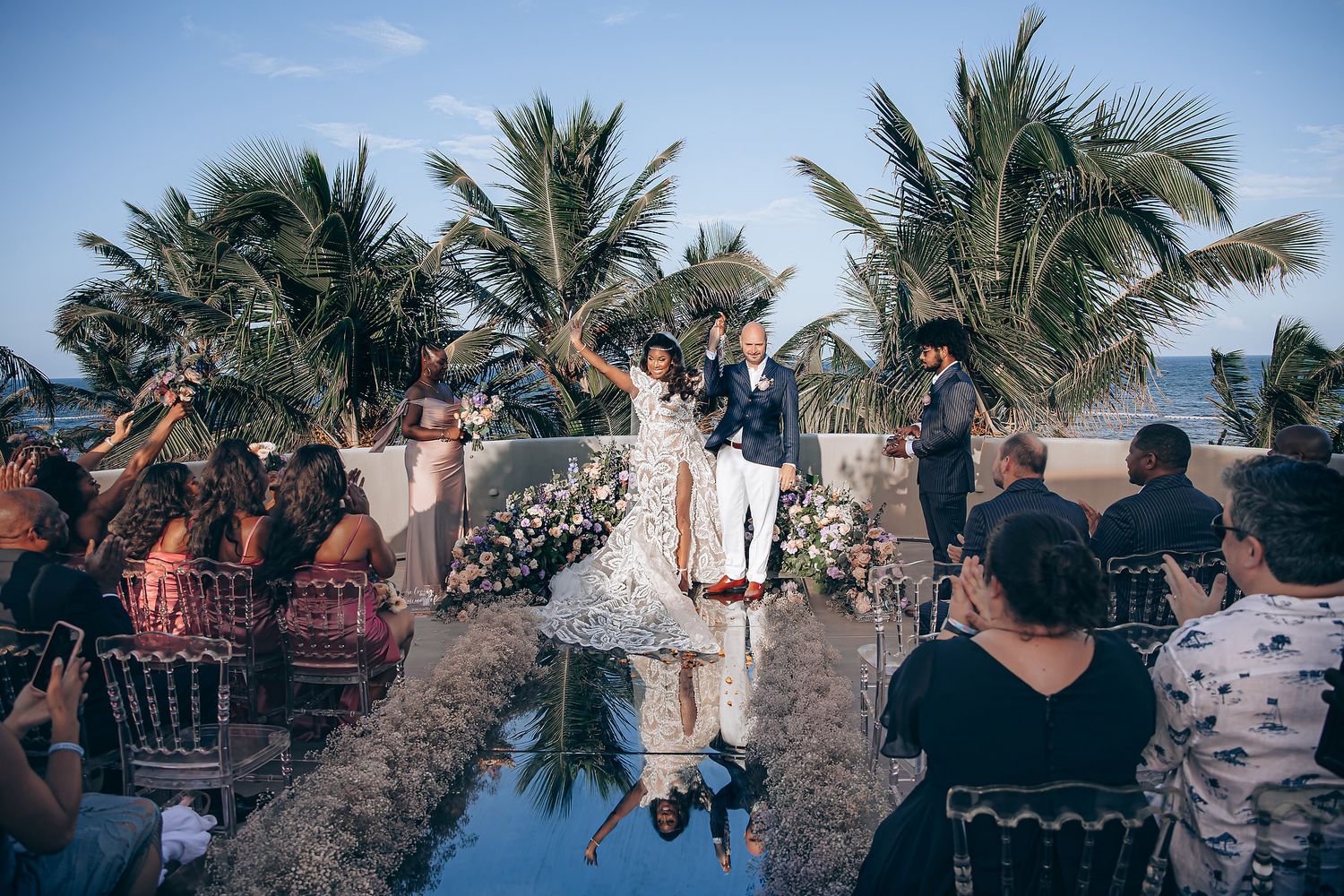 Tropical beach wedding ceremony with palm trees swaying in the breeze as guests gather under blue skies.