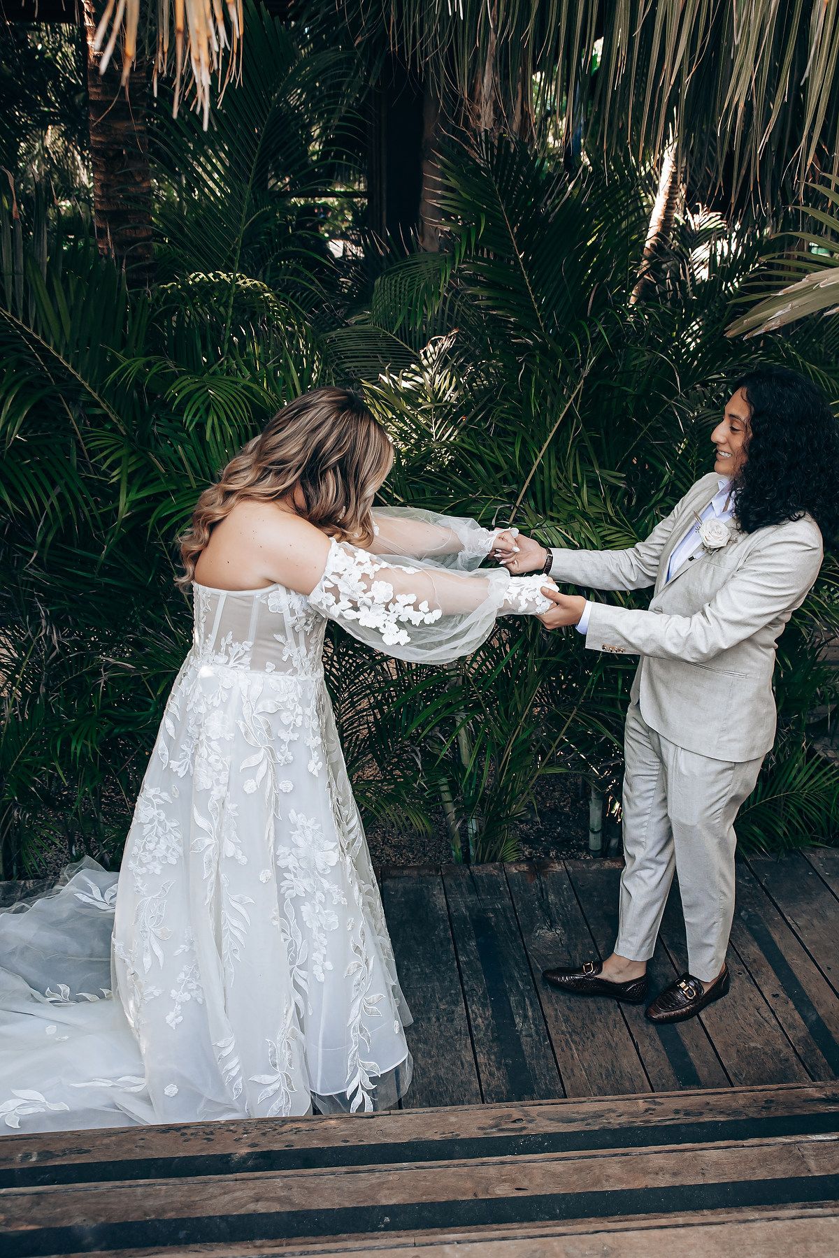 A couple in wedding attire shares a playful moment on a wooden deck surrounded by tropical plants.