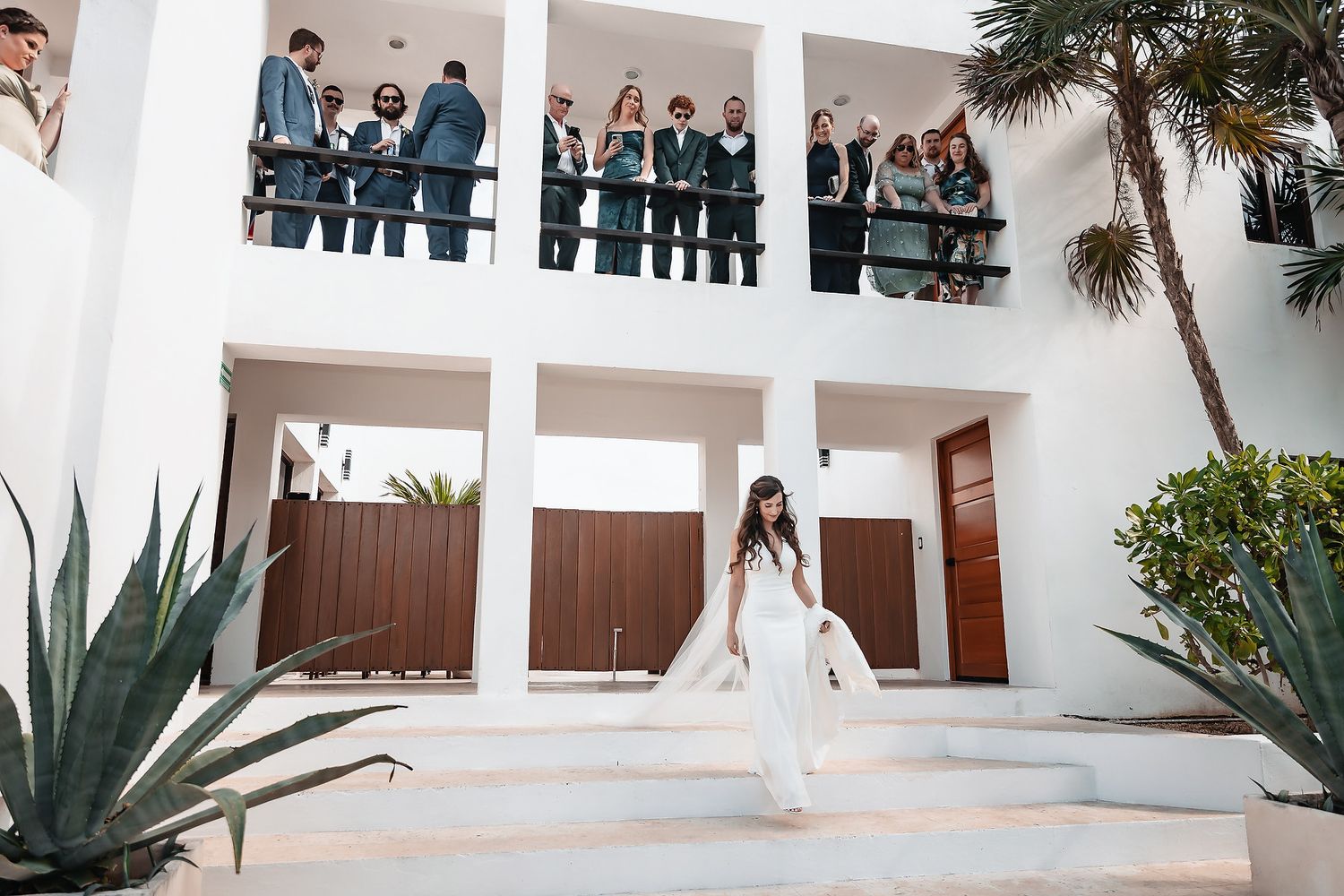 Bride in white gown descends steps of modern white villa while wedding guests watch from upper balcony.