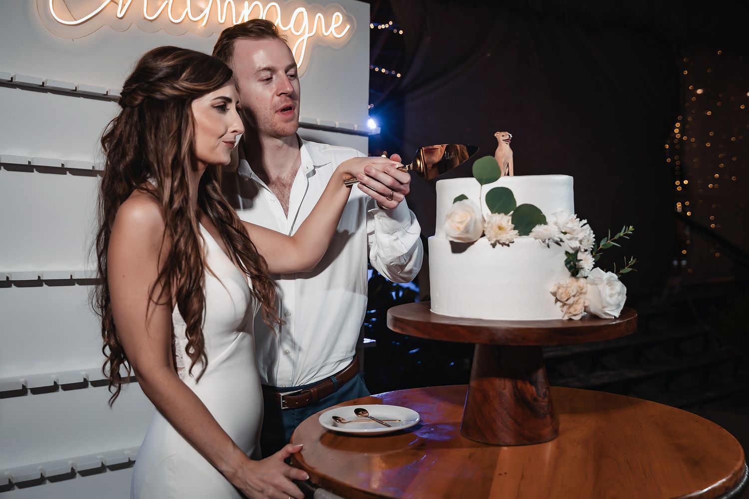 Bride and groom cutting their white floral wedding cake together at a romantic reception with neon sign backdrop.