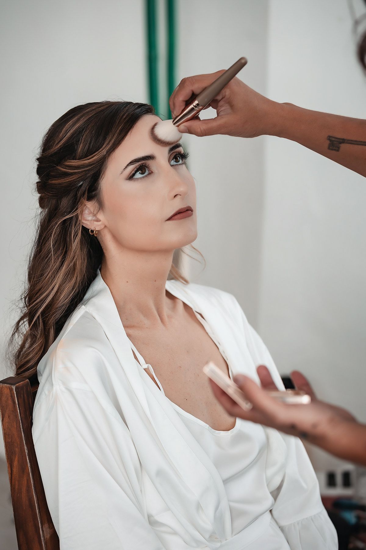 A makeup artist applies foundation to a brunette woman in a white robe sitting in a chair during bridal preparation.