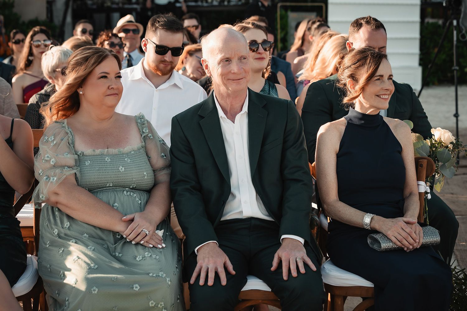 Three wedding guests seated outdoors, smiling during a ceremony, dressed in elegant attire at a sunny outdoor venue.