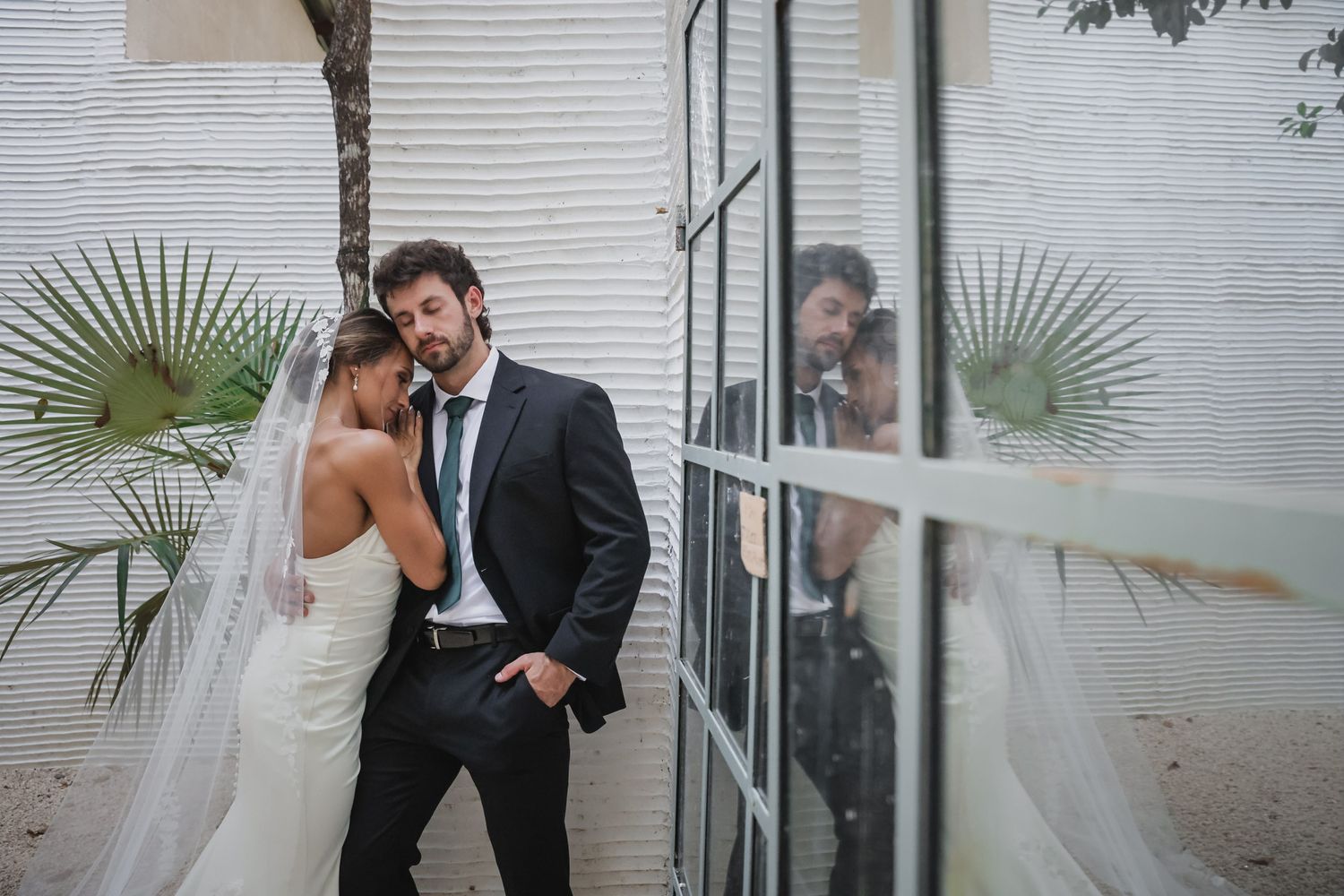 Bride and groom share an intimate moment against a white wall, with photographer reflected in glass door beside them.