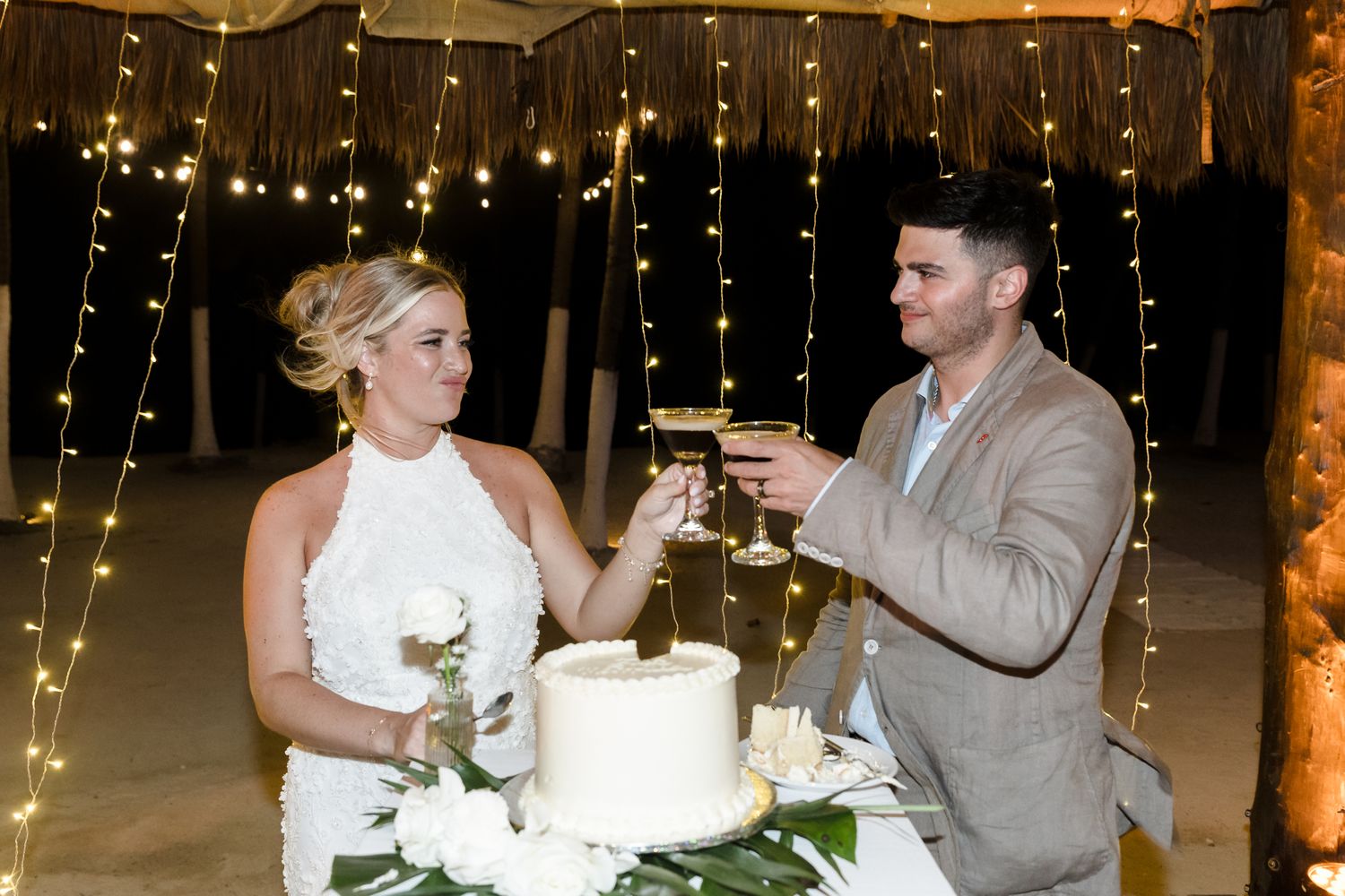 Bride and groom toasting with wine glasses beside a white wedding cake, surrounded by warm fairy lights backdrop.