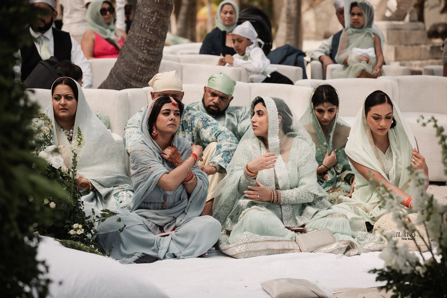 Guests in elegant mint green and blue dupattas sit on white cushions during an outdoor Sikh wedding ceremony.