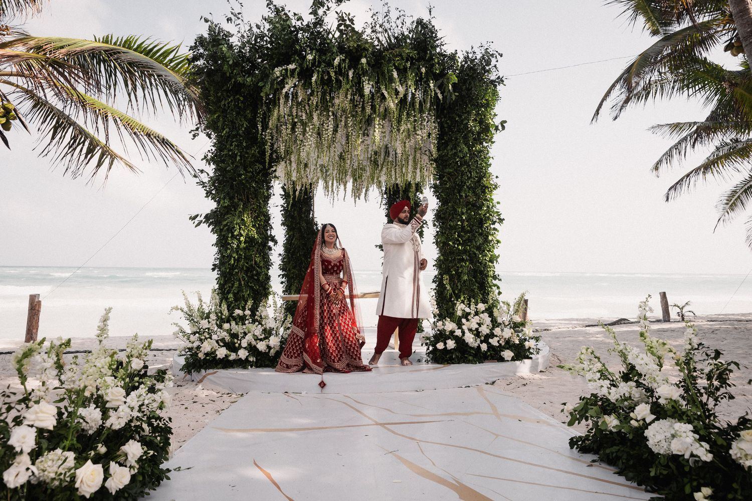 Indian bride and groom in traditional red and white wedding attire standing before greenery arch with white flowers on beach.