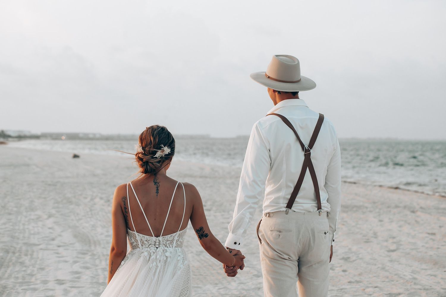 Couple walking hand in hand along a serene beach shoreline in elegant wedding attire.