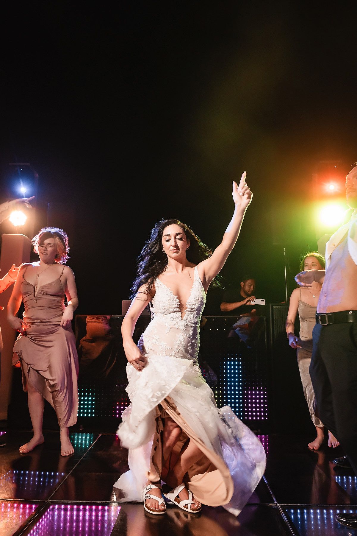 Bride in white gown dancing joyfully on illuminated dance floor at wedding reception with guests.