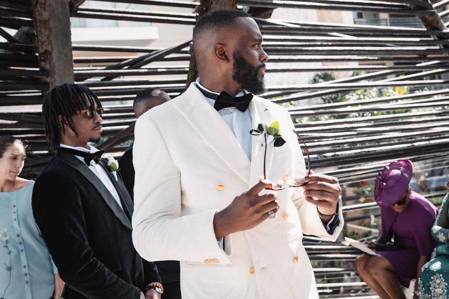 Groom in white double-breasted tuxedo holding rings at outdoor wedding ceremony.