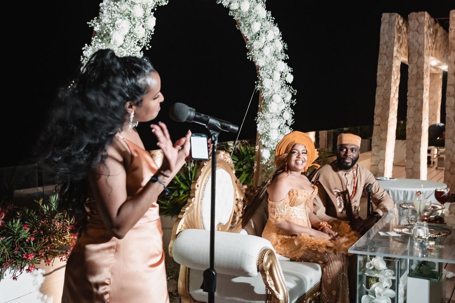 Woman giving speech at microphone during elegant outdoor African wedding reception ceremony at night.