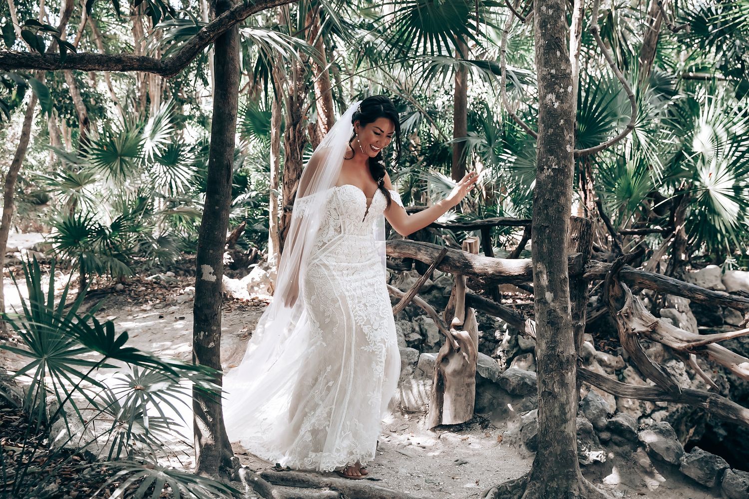 A person in a flowing white wedding dress walks through a lush tropical garden setting with palm trees and greenery.