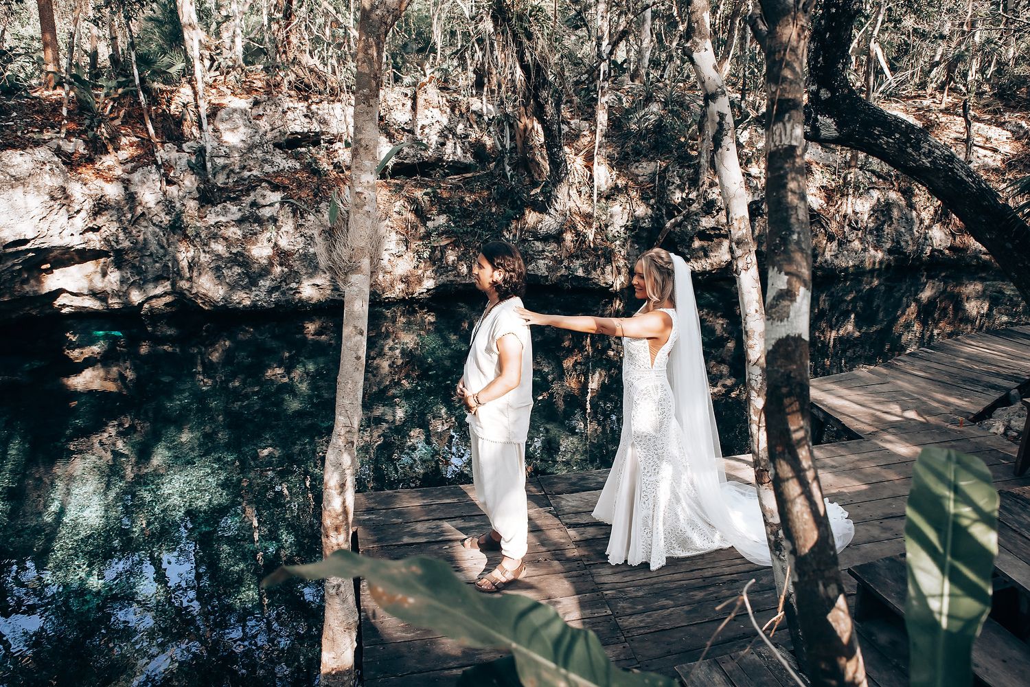 Wedding couple walks hand in hand along a rustic wooden pathway with trees and water in the background.