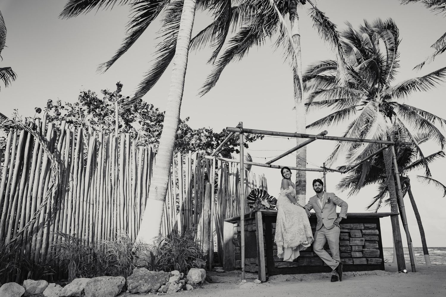 Black and white photo of wedding party relaxing near bamboo fence on beach.