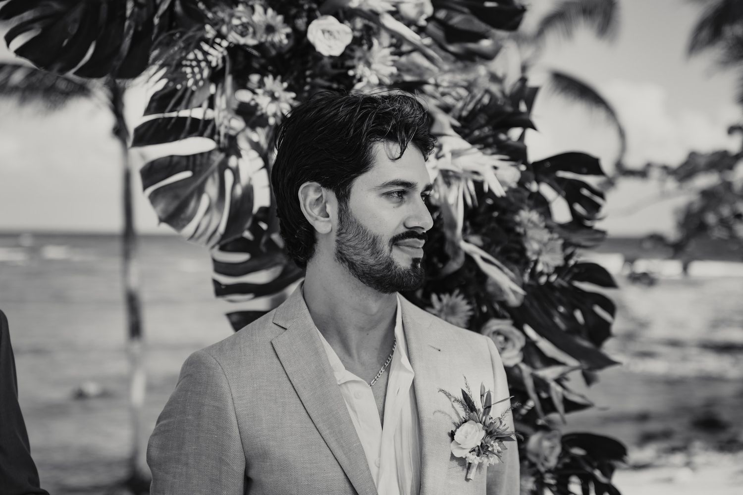 Emotional groom in light gray suit with boutonniere at tropical beach wedding ceremony.