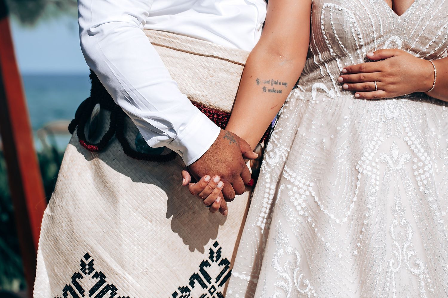 Close-up of two people holding hands while wearing white lace and embroidered dresses at an outdoor event.