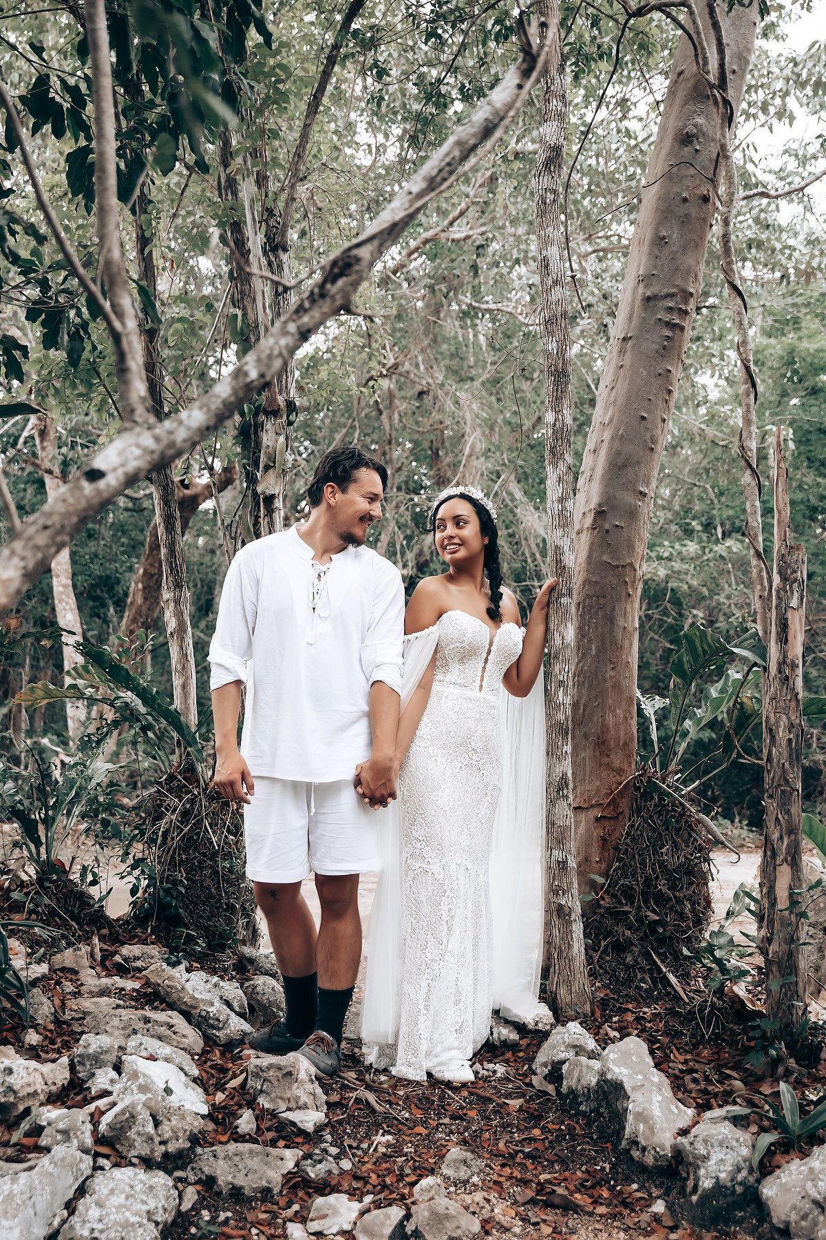A couple in white attire poses together in a wooded area with stone formations for their wedding photos.