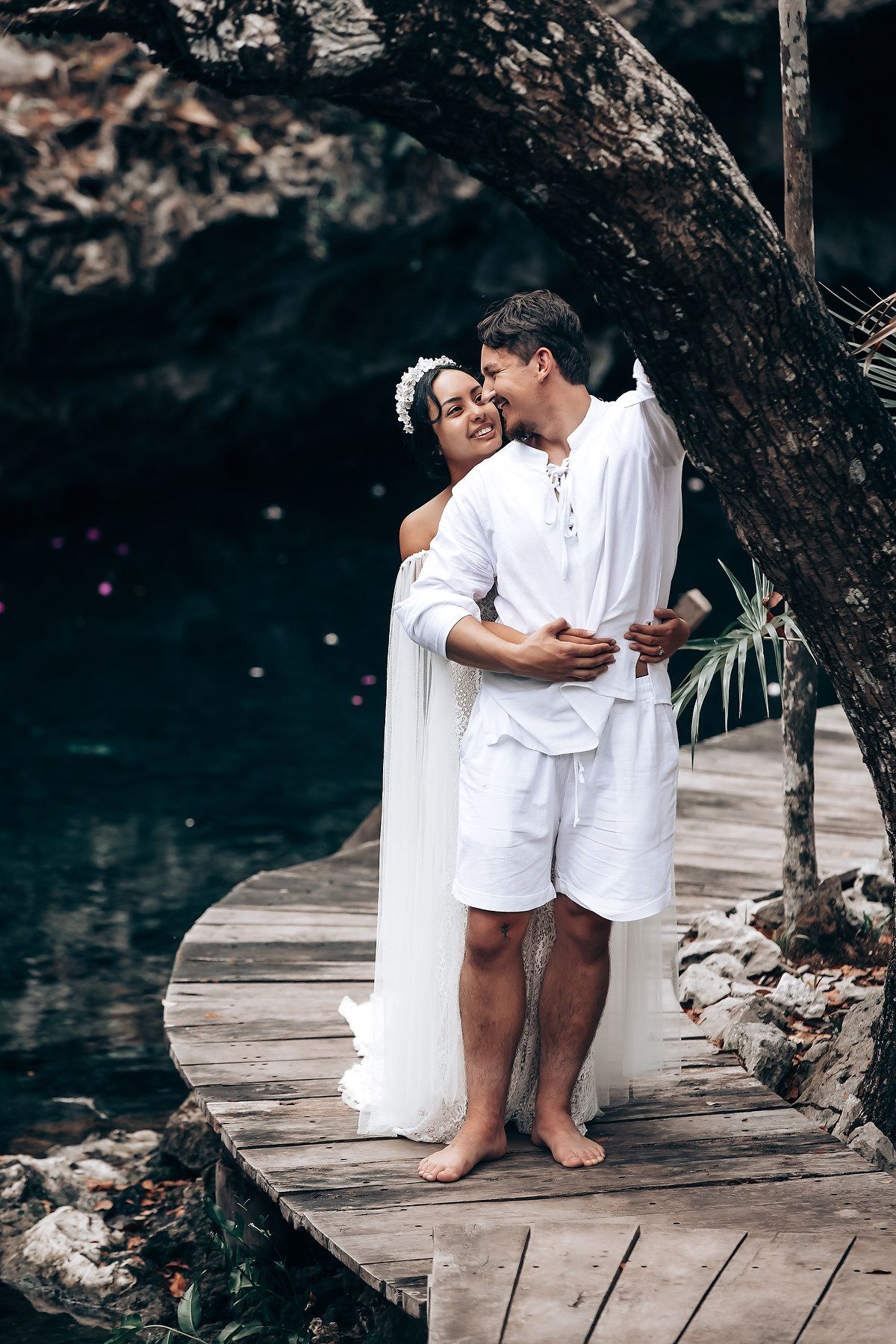 A couple in white clothing shares a tender moment while standing on a wooden boardwalk near water.