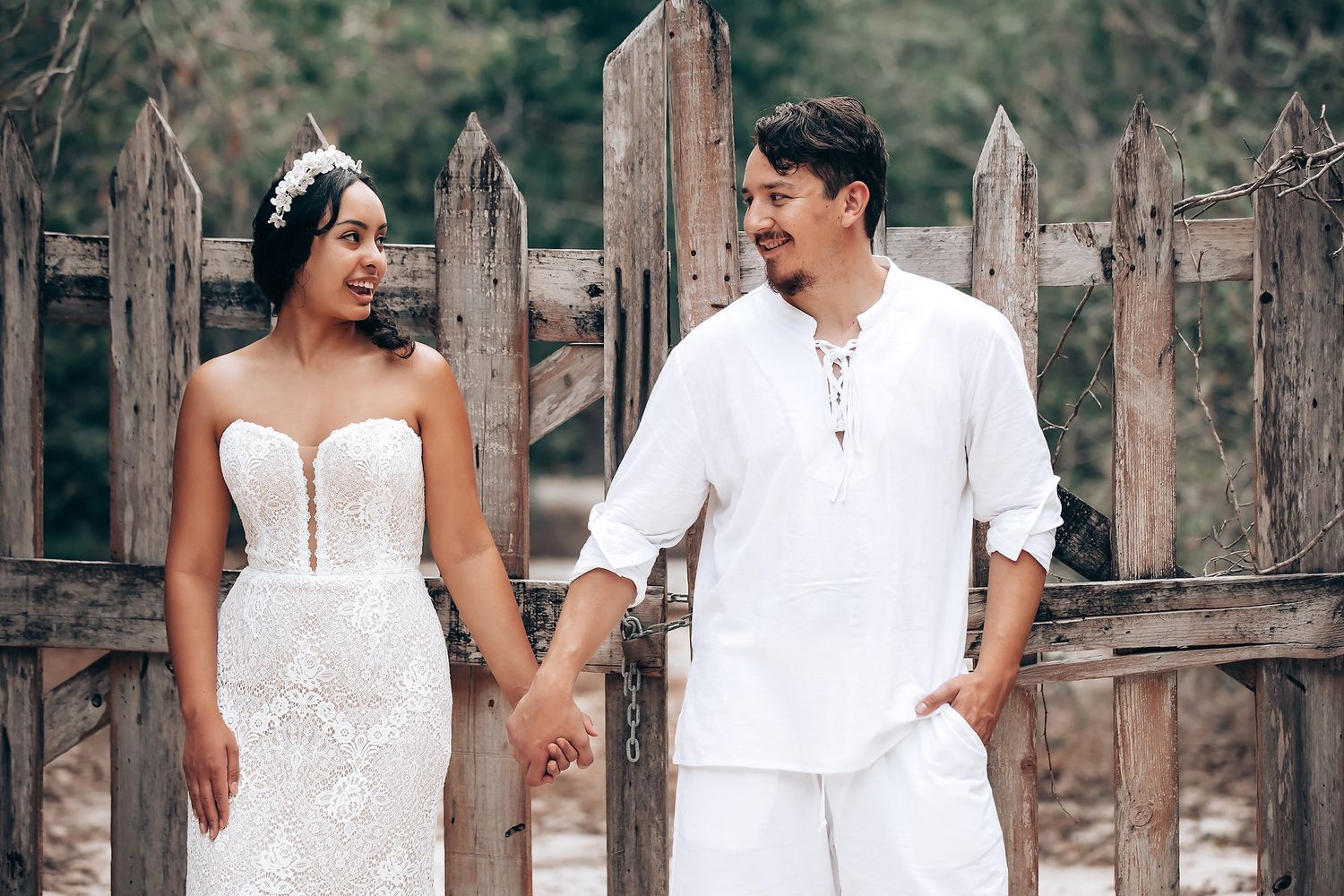 Couple in white outfits poses together in a rustic outdoor setting with wooden fence and trees in background.