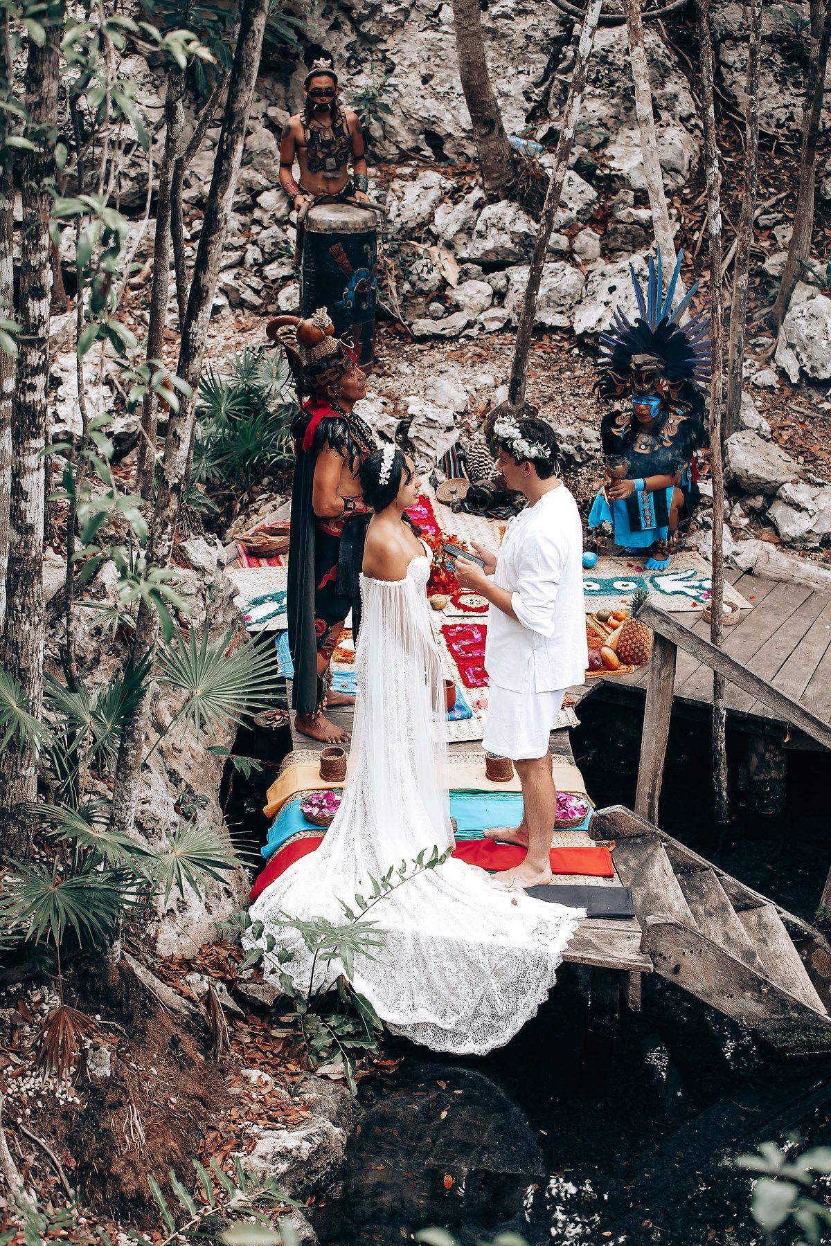 A traditional Mayan wedding ceremony takes place on a wooden platform over water in a natural cenote cave setting.