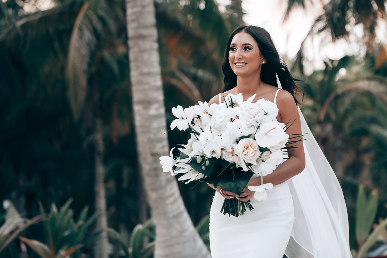 Bride holding elegant white floral bouquet against tropical palm background.