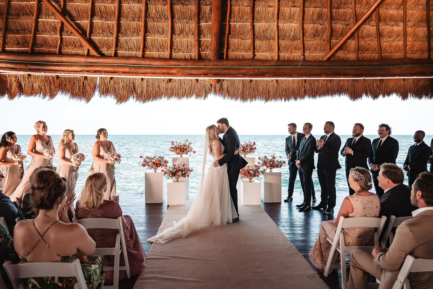 Wedding guests gather under a rustic thatched-roof palapa venue during a beach celebration.