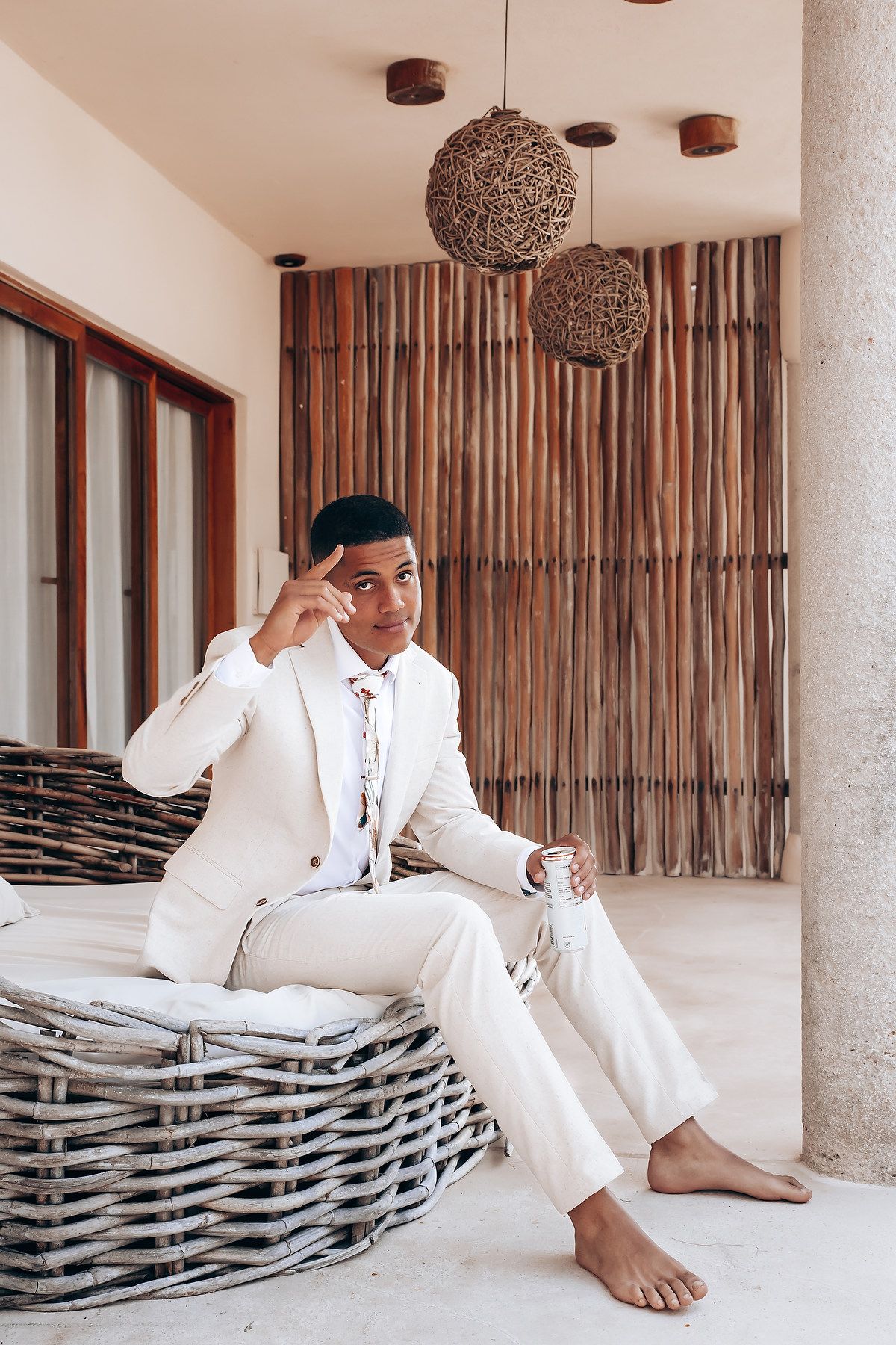 Stylish groom relaxes on woven outdoor furniture in a crisp white suit.