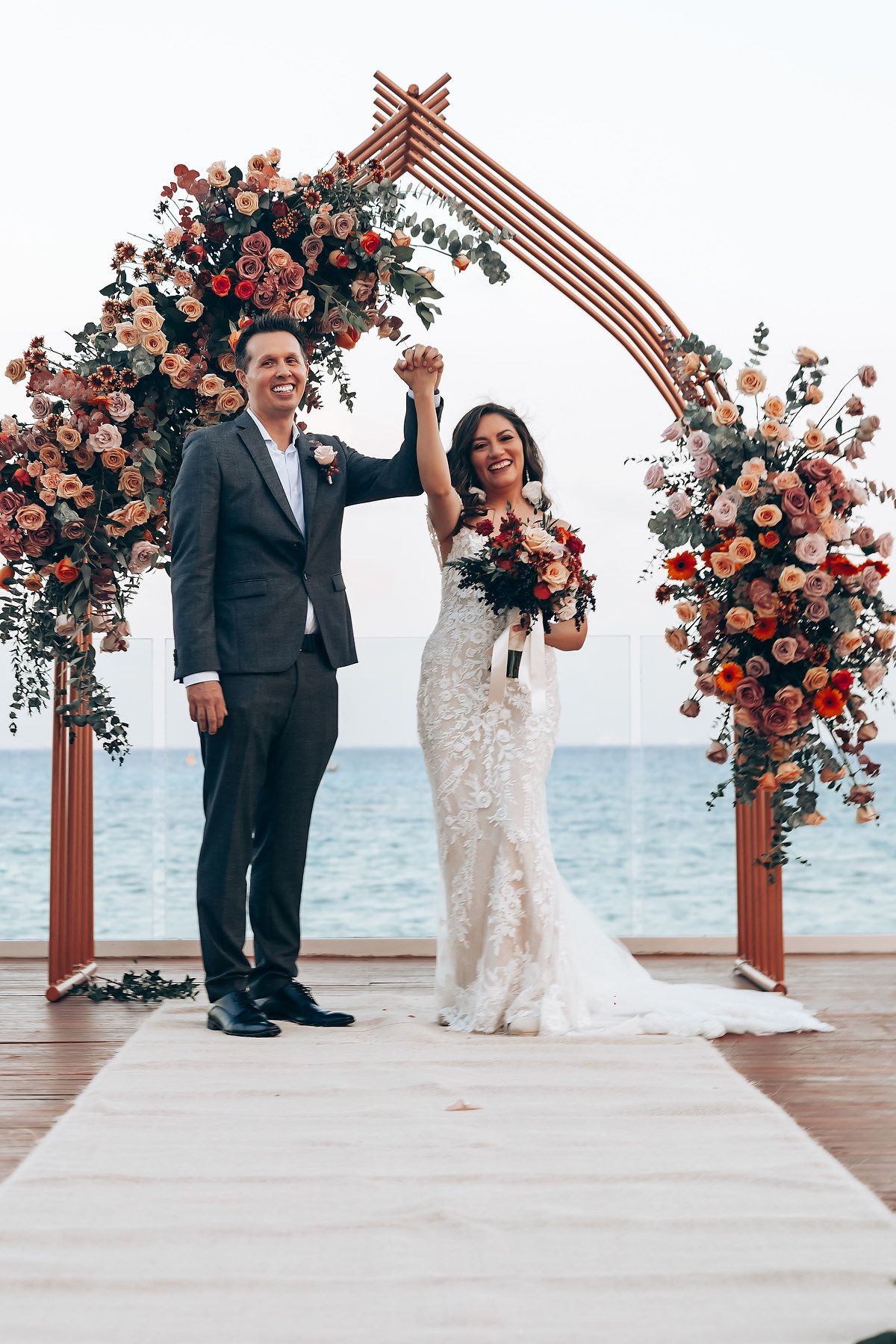 Newlyweds celebrate under a wooden arch adorned with coral and pink flowers against an ocean backdrop.