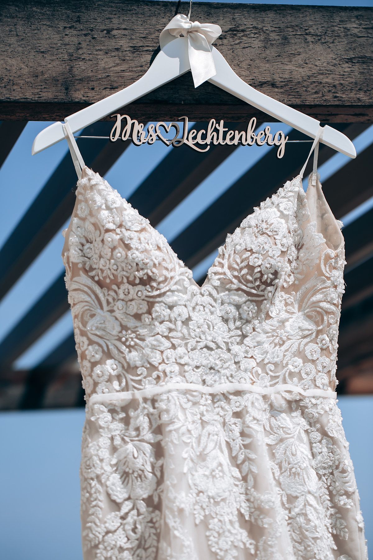 Elegant white lace wedding dress with intricate floral details hanging on display against rustic wooden wall.