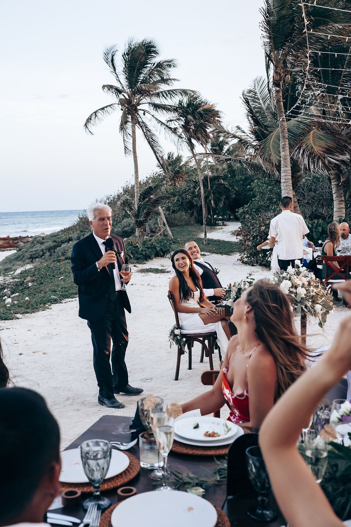 Wedding reception dinner taking place on a beachfront patio with palm trees at sunset.