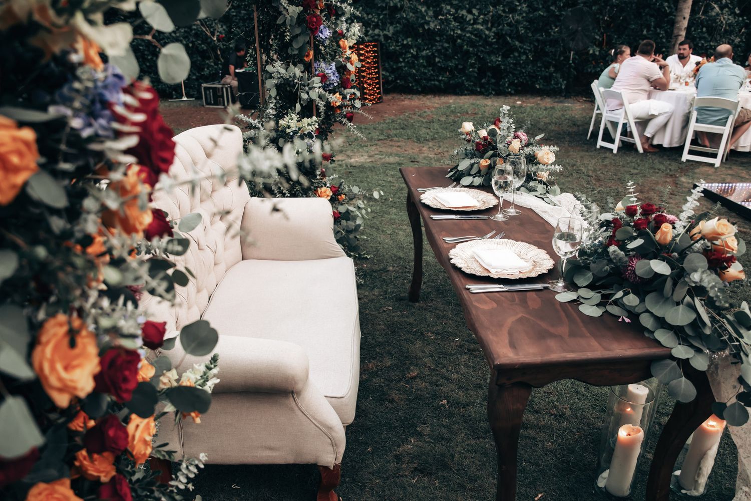 Elegant outdoor wedding reception with floral arrangements, white sofa, wooden table settings, and guests dining in background.