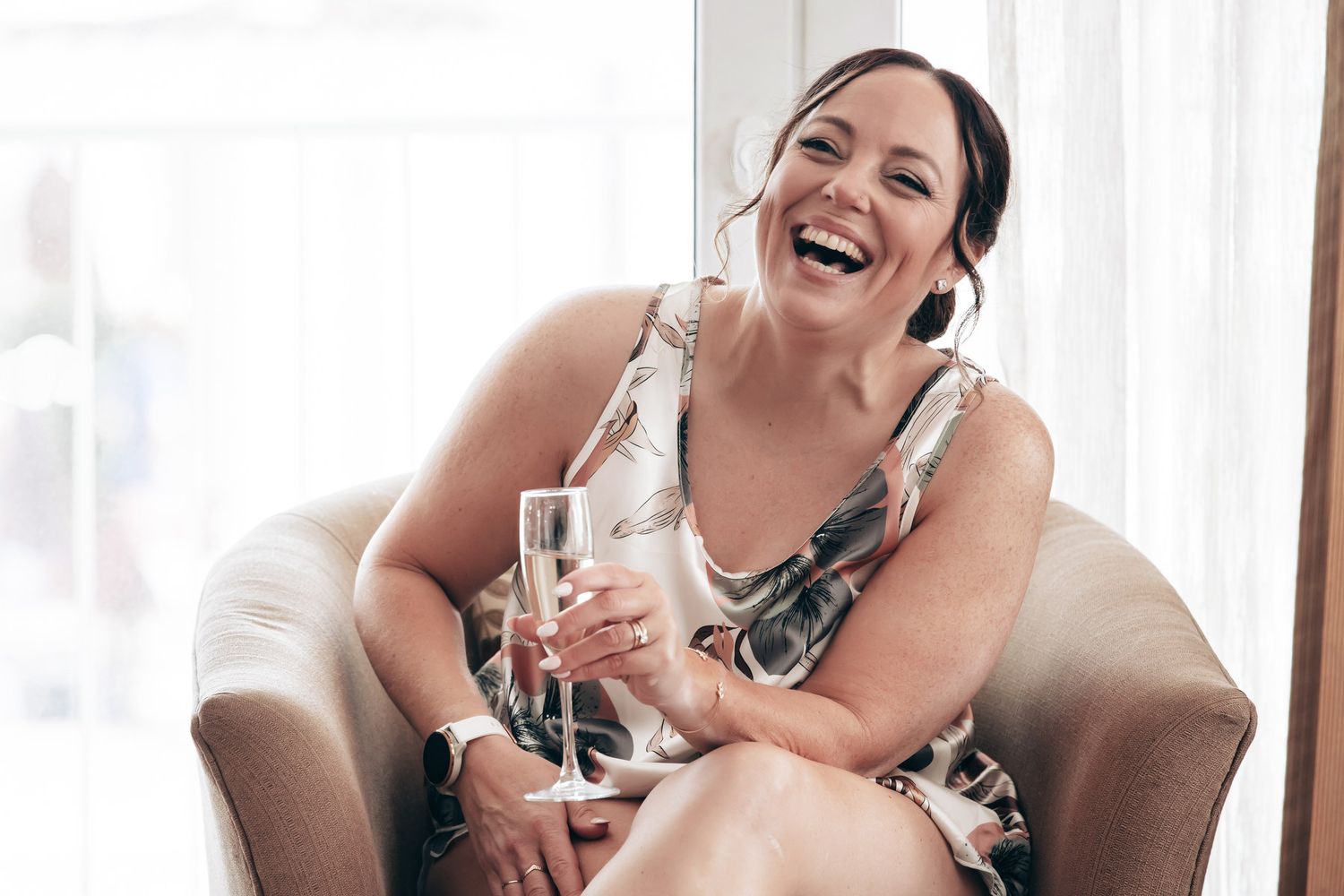 A laughing woman in a floral dress holds a champagne glass while relaxing in a chair at a wedding celebration.