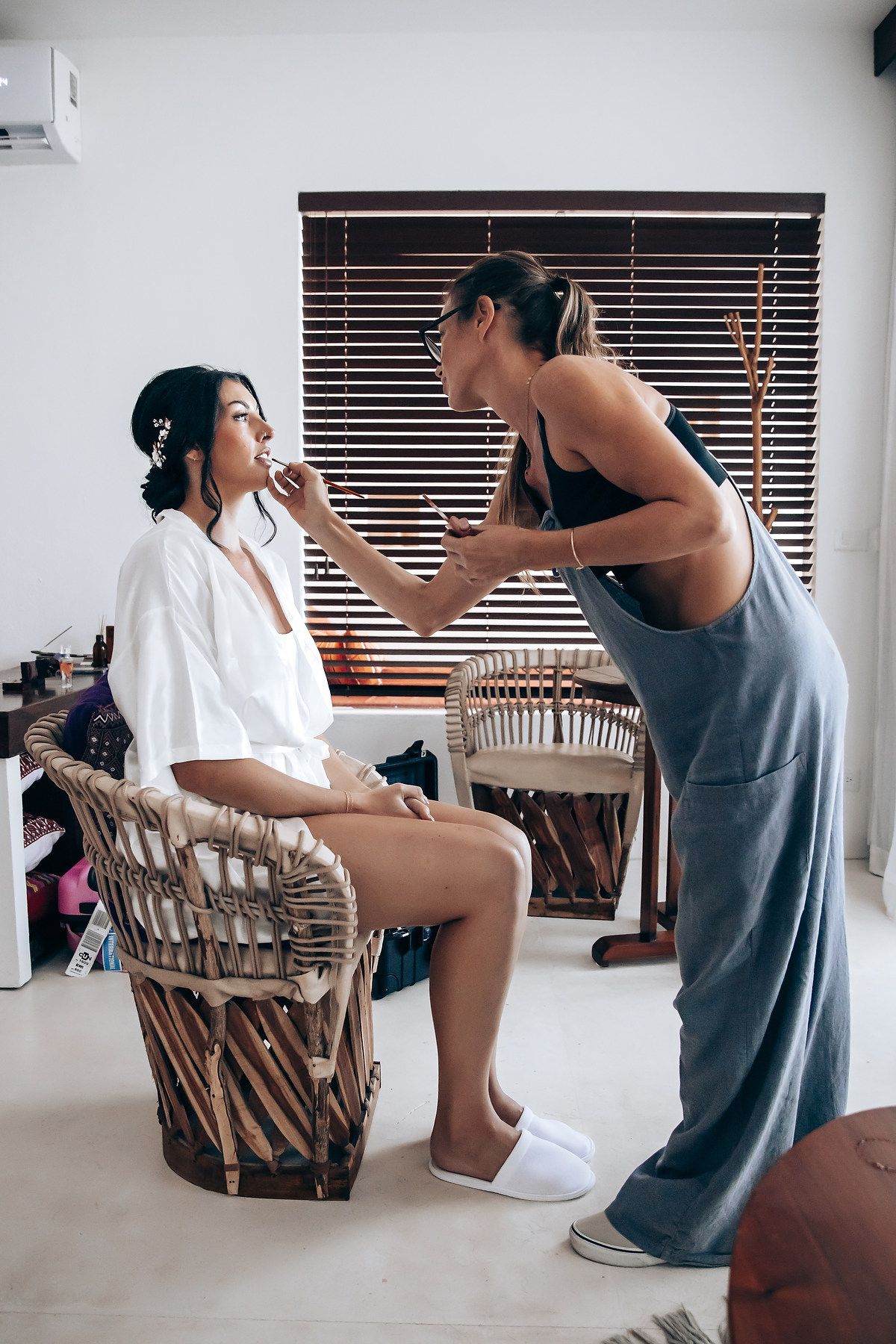 Two women interact in a room with a window and wooden blinds.