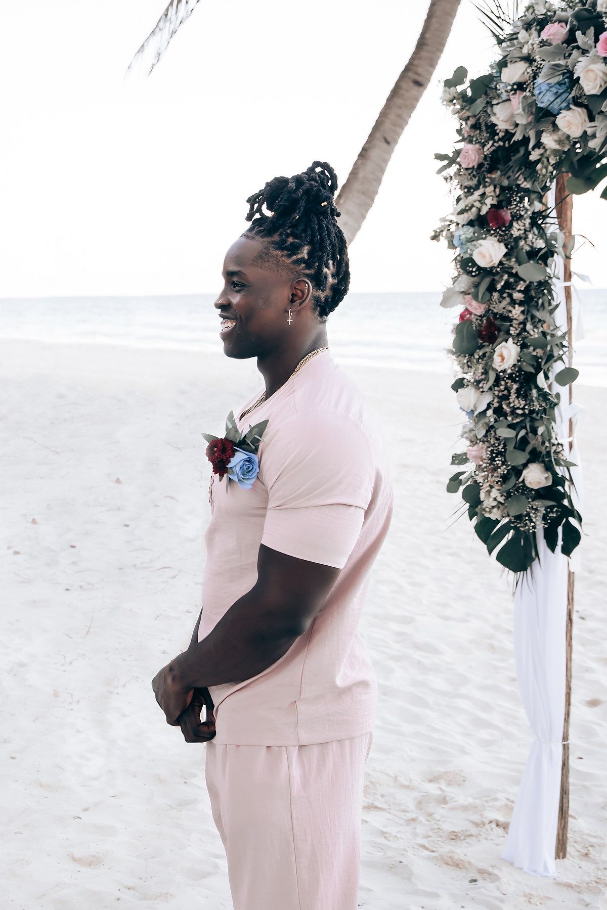 A woman in a pink dress stands in front of a floral arch on a beach.