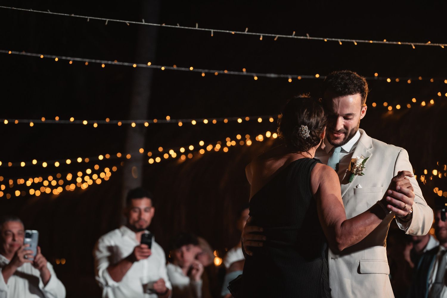 Couple shares romantic first dance under warm string lights at outdoor wedding reception at night.