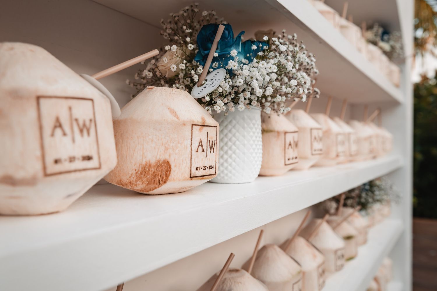 Personalized coconut drinks with initials displayed on white shelves at tropical wedding.