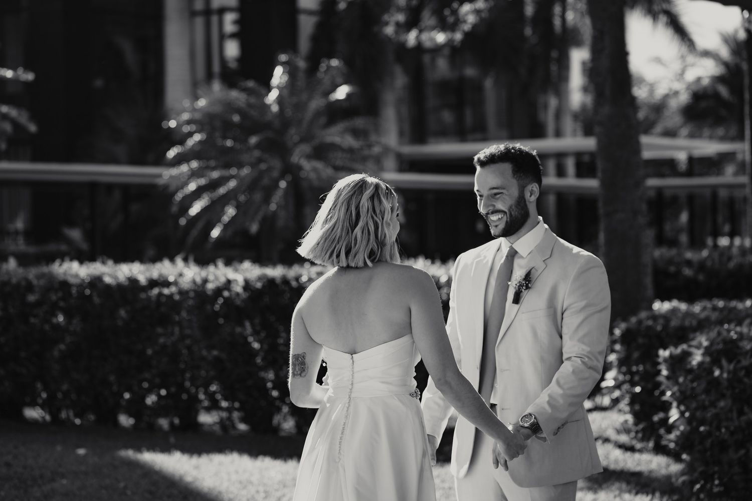 Black and white photo of smiling groom in suit holding hands with bride outdoors near palm trees.