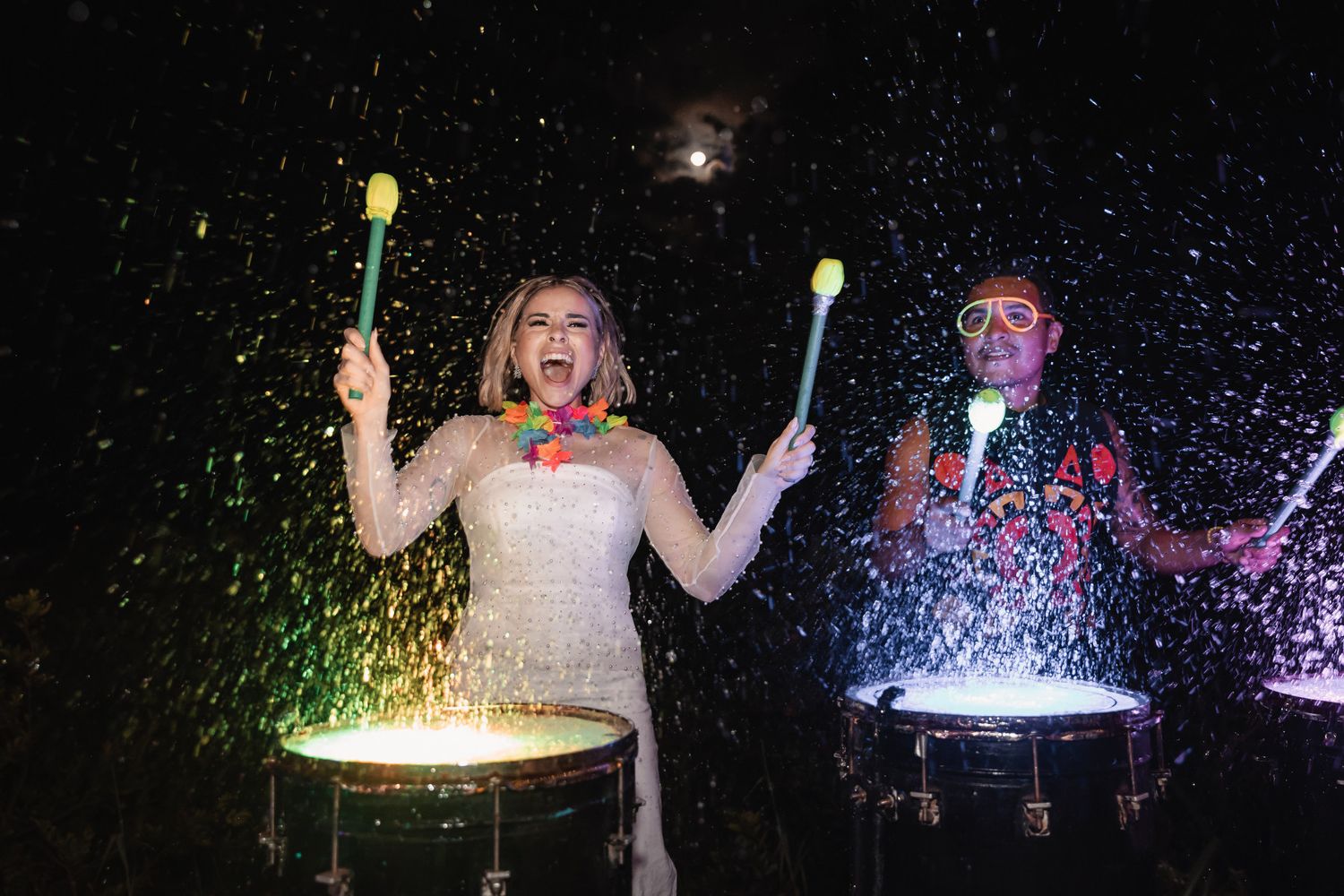 Two women in wedding attire joyfully playing illuminated drums outdoors at night with colorful drumsticks.