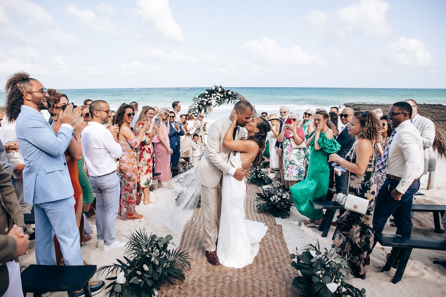 Wedding guests gather on a sunny beach with ocean waves in the background during an outdoor ceremony.