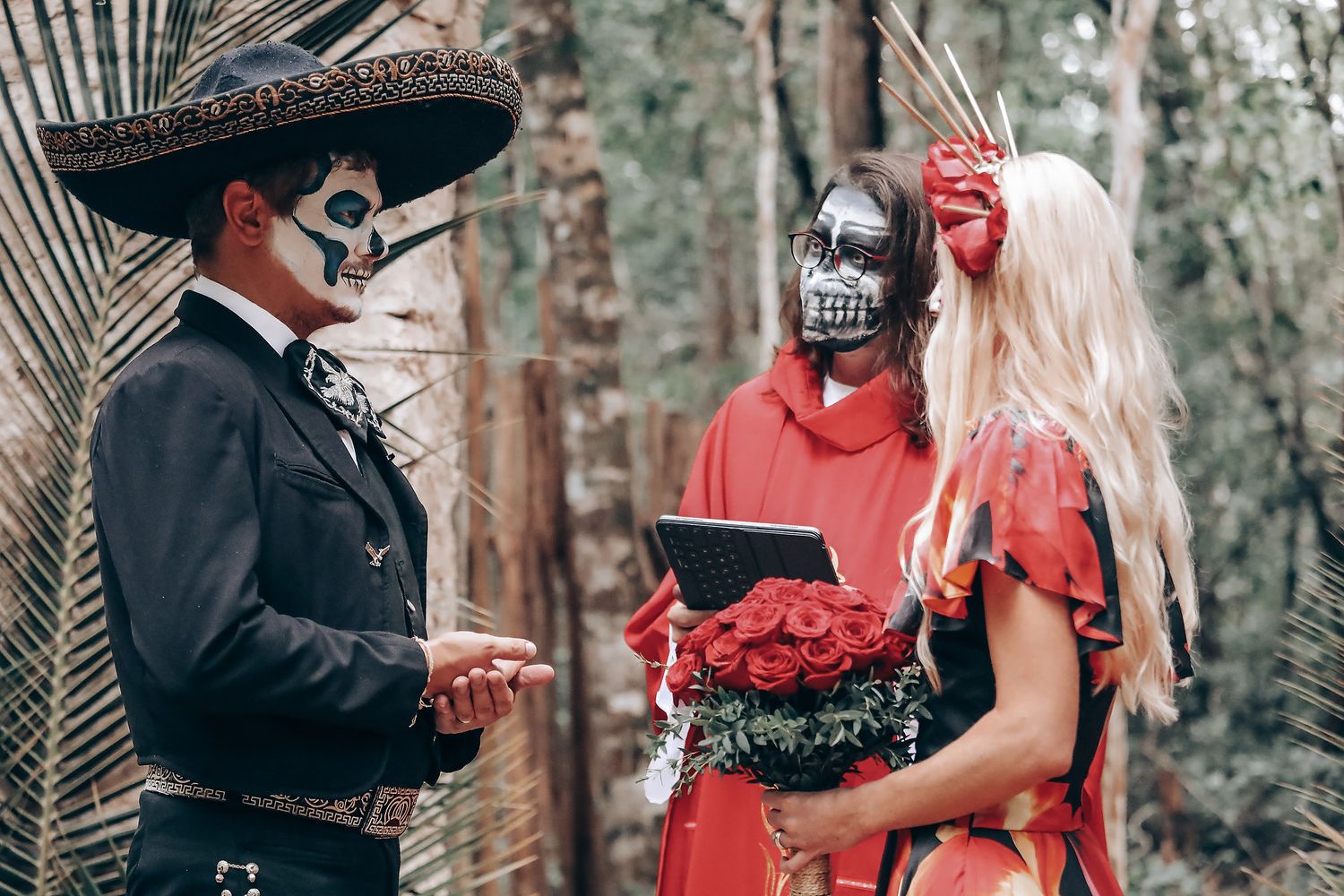 A couple in Day of the Dead costumes share an intimate moment in a wooded setting with stone walls and palm fronds.